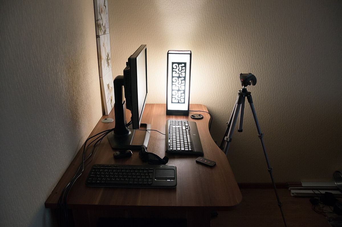 Pracovný stôl s monitorom na ramene, klávesnicou a lampou v tlmenom svetle